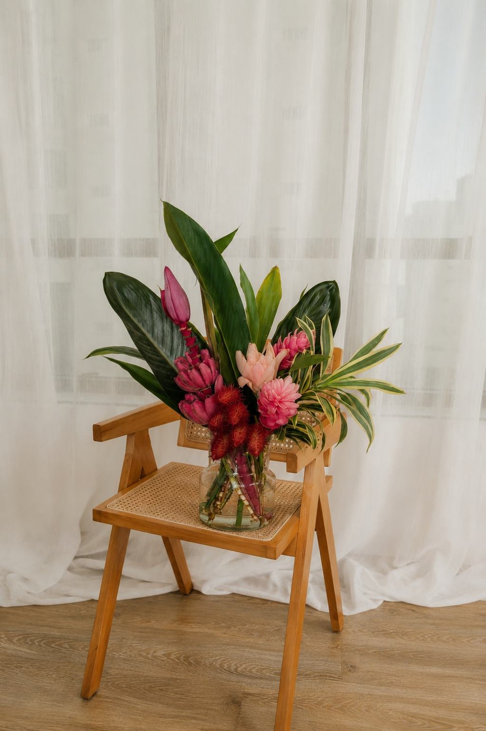 Arranjo tropical de flores sobre uma cadeira de madeira, com cortina branca ao fundo